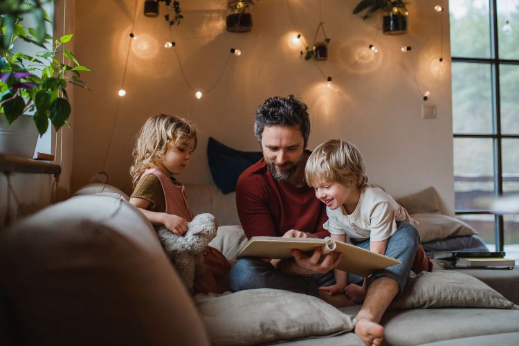 Vater sitzt mit zwei Kindern auf einem gemütlichen Sofa und liest ihnen ein Buch vor, in einem warmen, dekorativ beleuchteten Wohnzimmer mit Pflanzen.